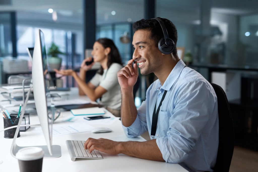 Local helpdesk technician ready to assist — Divine Logic Clean-shaven support technician in dress shirt and tie answers a call at a dual-monitor desk.